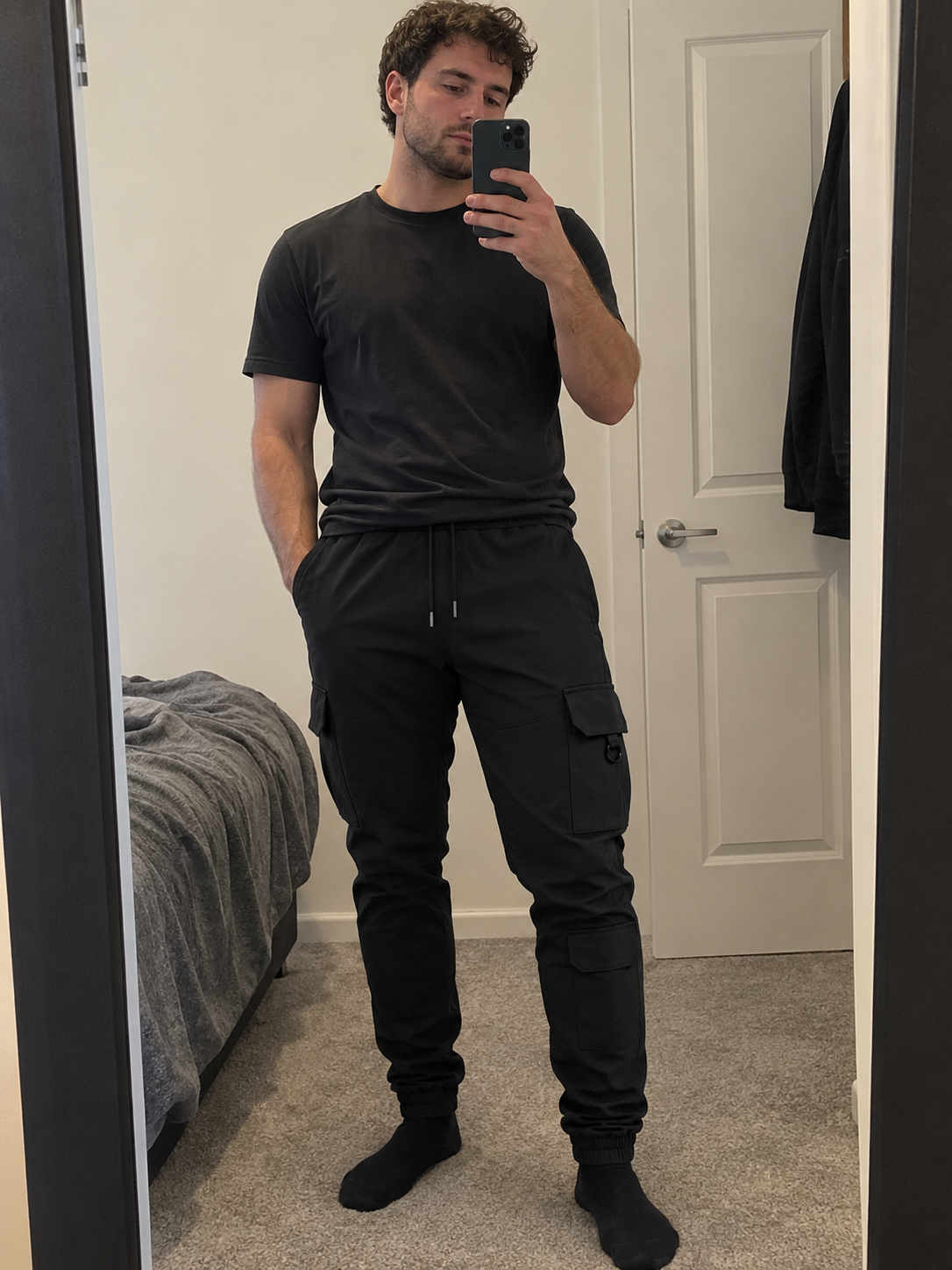 A man takes a mirror selfie in a bedroom wearing a black crewneck t-shirt and black cargo jogger pants with black socks.