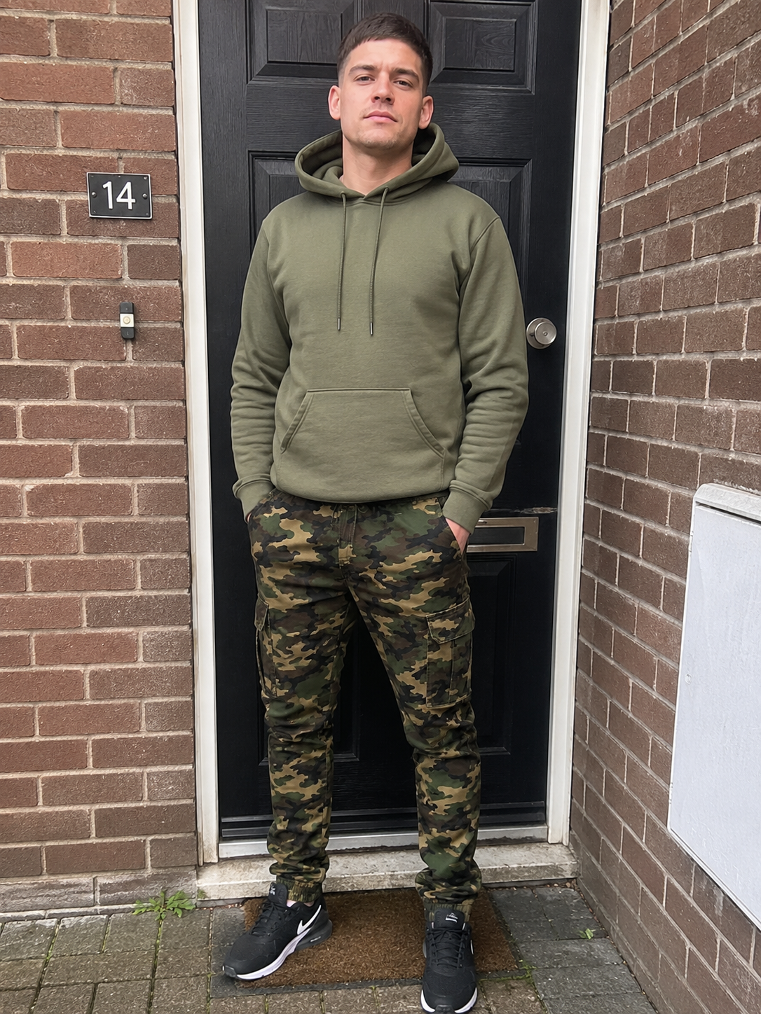 A man standing in front of a black door set in a brick wall, wearing an olive green hoodie and camouflage jogger pants.