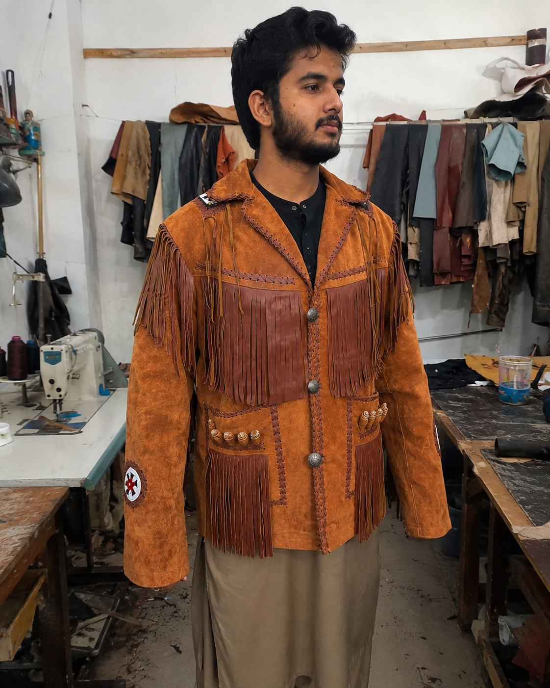A man wearing a handmade tan suede and leather fringe jacket with silver buttons, standing in a workshop with sewing machines and other garments in the background.